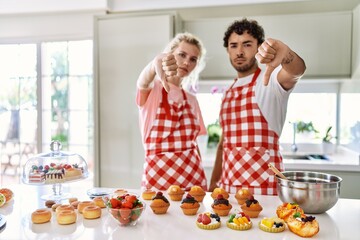 Couple of wife and husband cooking pastries at the kitchen looking unhappy and angry showing rejection and negative with thumbs down gesture. bad expression.
