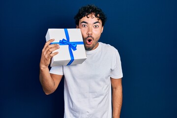 Young hispanic man holding gift scared and amazed with open mouth for surprise, disbelief face