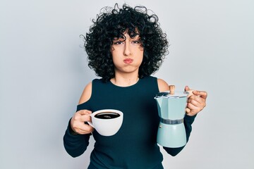 Young middle east woman holding italian coffee maker puffing cheeks with funny face. mouth inflated...