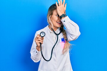 Beautiful young blonde doctor woman holding stethoscope surprised with hand on head for mistake,...