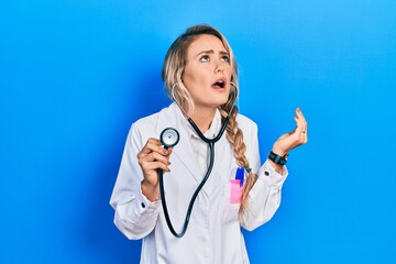 Beautiful young blonde doctor woman holding stethoscope crazy and mad shouting and yelling with aggressive expression and arms raised. frustration concept.