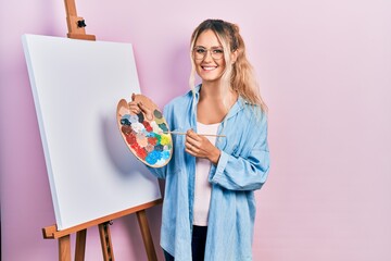 Beautiful young blonde woman standing drawing with palette by painter easel stand smiling with a happy and cool smile on face. showing teeth.