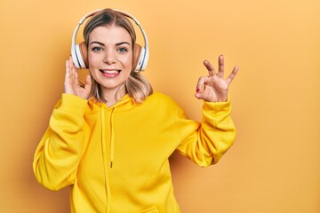 Beautiful caucasian woman listening to music using headphones doing ok sign with fingers, smiling friendly gesturing excellent symbol