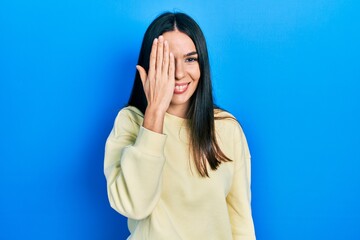 Young brunette woman wearing casual sweatshirt covering one eye with hand, confident smile on face and surprise emotion.