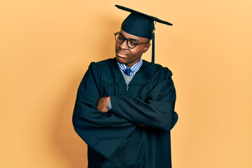 Young african american man wearing graduation cap and ceremony robe looking to the side with arms...