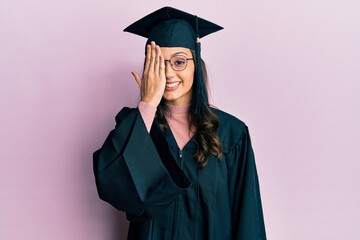 Young hispanic woman wearing graduation cap and ceremony robe covering one eye with hand, confident...