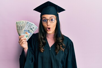 Young hispanic woman wearing graduation uniform holding usa dollars scared and amazed with open...