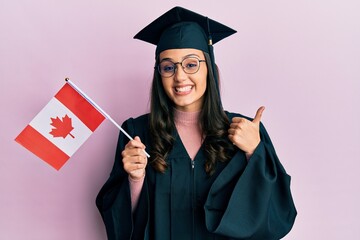 Young hispanic woman wearing graduation uniform holding canada flag smiling happy and positive,...