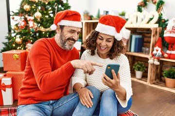 Middle age hispanic couple smiling happy wearing christmas hat. Sitting on the floor using smartphone at home.