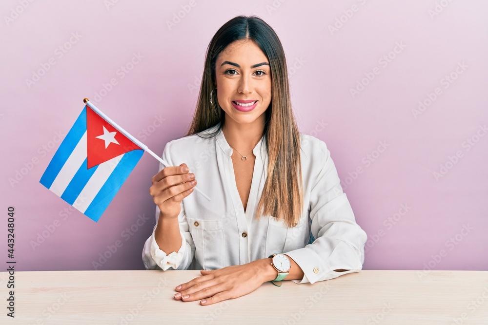 Wall mural young hispanic woman holding cuba flag sitting on the table looking positive and happy standing and 