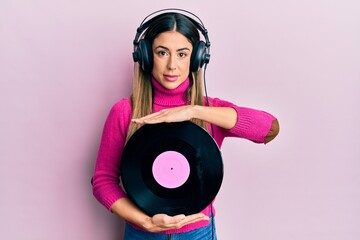 Young hispanic woman listening to music using headphones holding vinyl disc relaxed with serious...