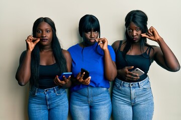 Three young african american friends using smartphone mouth and lips shut as zip with fingers....
