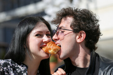 Happy young couple in morning and having breakfast. Cheerful man and woman bite a croissant at the same time.