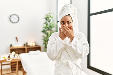 Young brunette woman wearing towel and bathrobe standing at beauty center shocked covering mouth with hands for mistake. secret concept.