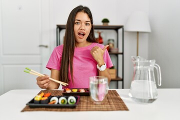 Obraz na płótnie Canvas Young brunette woman eating sushi using chopsticks surprised pointing with hand finger to the side, open mouth amazed expression.