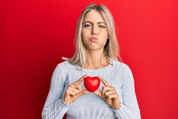 Beautiful blonde woman holding heart puffing cheeks with funny face. mouth inflated with air, catching air.