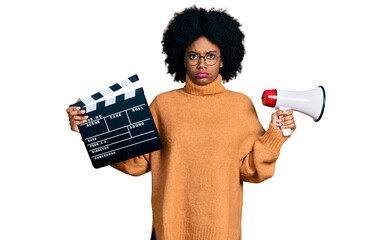 Young african american woman holding video film clapboard and megaphone skeptic and nervous,...