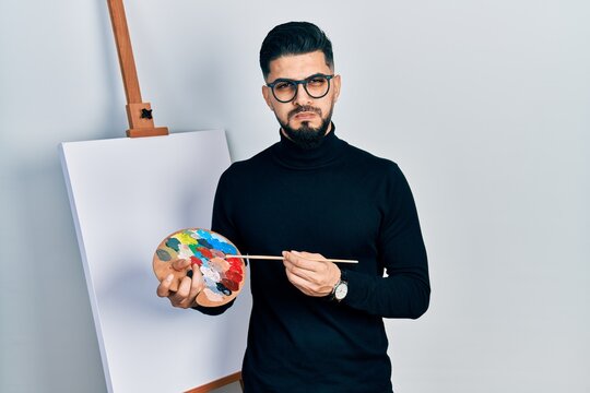 Handsome man with beard holding palette and brush close to easel stand puffing cheeks with funny face. mouth inflated with air, catching air.