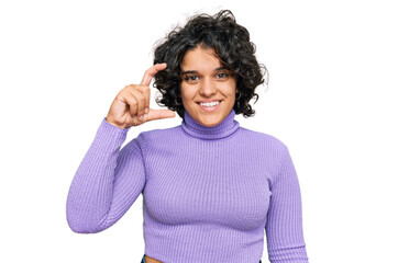 Young hispanic woman with curly hair wearing casual clothes smiling and confident gesturing with hand doing small size sign with fingers looking and the camera. measure concept.