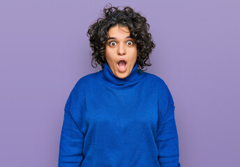 Young hispanic woman with curly hair wearing turtleneck sweater afraid and shocked with surprise and amazed expression, fear and excited face.