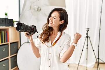 Beautiful caucasian woman working as photographer at photography studio very happy and excited doing winner gesture with arms raised, smiling and screaming for success. celebration concept. - obrazy, fototapety, plakaty