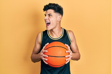 Young hispanic man holding basketball ball angry and mad screaming frustrated and furious, shouting...