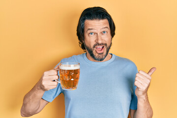 Middle age caucasian man drinking a jar of beer pointing thumb up to the side smiling happy with open mouth