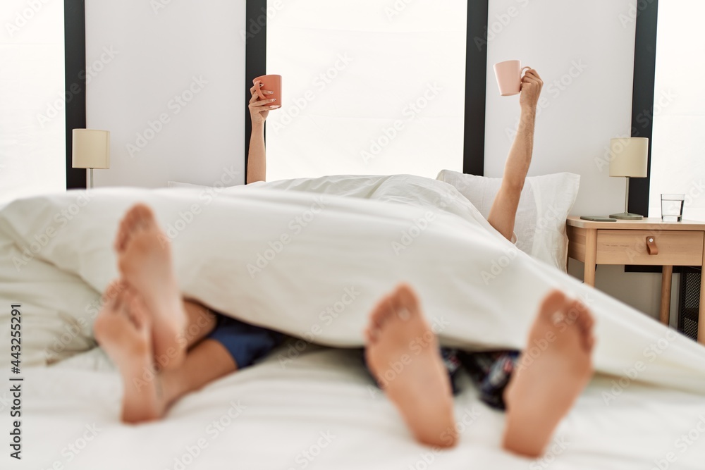 Poster couple awake under blanket. having breakfast holding cup of coffee raised up on the bed.