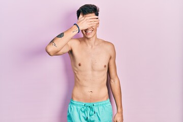 Fototapeta premium Young hispanic man wearing swimwear shirtless smiling and laughing with hand on face covering eyes for surprise. blind concept.