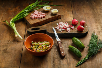 Okroshka in a clay plate on a table on a wooden table next to vegetables on boards and a knife.