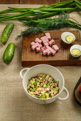 Okroshka in a soup bowl on a wooden table next to greens on planks and a knife.