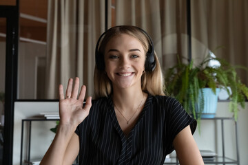 Head shot portrait of smiling businesswoman in headphones waving hand and looking at camera, business mentor coach teacher greeting students viewers, recording webinar, employee making video call