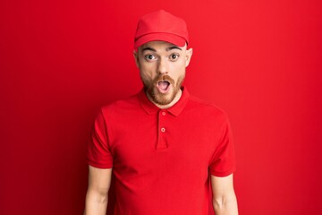 Young redhead man wearing delivery uniform and cap scared and amazed with open mouth for surprise, disbelief face