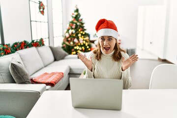 Middle age woman wearing santa claus hat using laptop crazy and mad shouting and yelling with aggressive expression and arms raised. frustration concept.