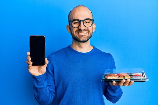 Young hispanic man holding take away food showing smartphone screen smiling with a happy and cool smile on face. showing teeth.