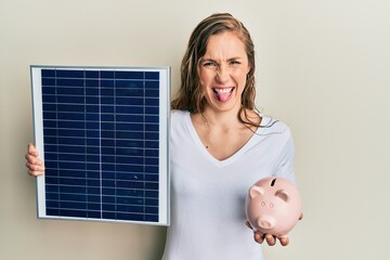 Young blonde woman holding photovoltaic solar panel and piggy bank sticking tongue out happy with...
