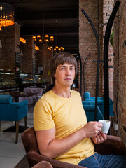 Fototapeta premium Man drinks coffee in cafe. Loft designed interior with red brick walls and plush chairs. Breakfast and morning cup of coffee away from home.
