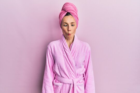 Young blonde woman wearing shower towel cap and bathrobe making fish face with lips, crazy and comical gesture. funny expression.