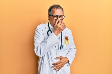 Middle age indian man wearing doctor coat and stethoscope smelling something stinky and disgusting, intolerable smell, holding breath with fingers on nose. bad smell
