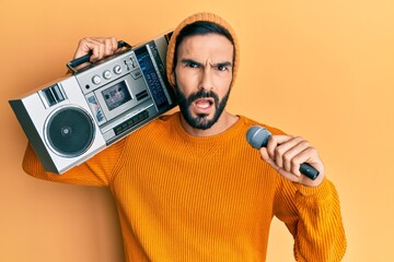 Young hispanic man holding boombox, listening to music singing with microphone in shock face,...
