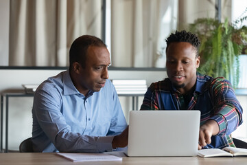 Two African American employees working on project together, using laptop pointing at screen,...