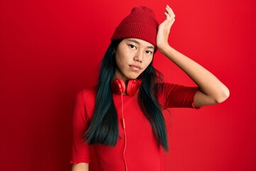 Young chinese woman listening to music using headphones surprised with hand on head for mistake,...