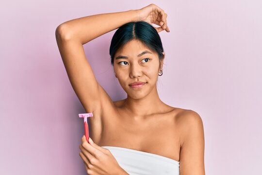 Young chinese woman wearing shower towel using razor smiling looking to the side and staring away thinking.