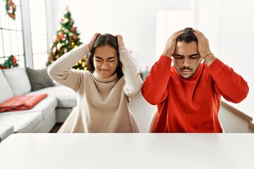 Young hispanic couple sitting on the table by christmas tree suffering from headache desperate and stressed because pain and migraine. hands on head.