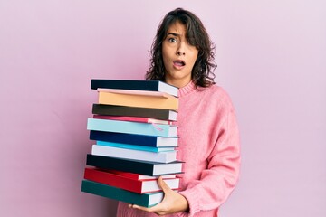 Young hispanic woman holding a pile of books in shock face, looking skeptical and sarcastic, surprised with open mouth