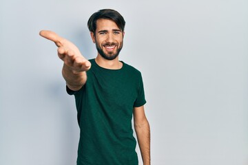 Fototapeta premium Young hispanic man wearing casual white tshirt smiling friendly offering handshake as greeting and welcoming. successful business.