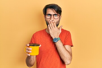 Young hispanic man drinking a cup of coffee covering mouth with hand, shocked and afraid for...