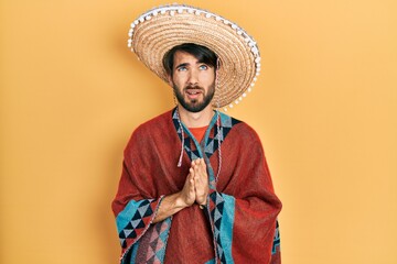 Young hispanic man holding mexican hat begging and praying with hands together with hope expression...
