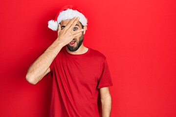Young hispanic man wearing christmas hat peeking in shock covering face and eyes with hand, looking through fingers with embarrassed expression.