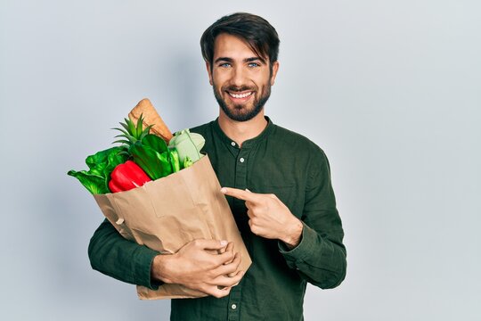 Young hispanic man holding paper bag with bread and groceries smiling happy pointing with hand and finger